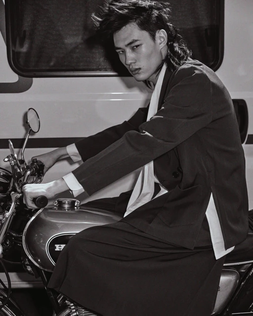 Monochrome shot of a young adult man in a suit sitting on a vintage Honda motorcycle, with a trailer visible in the background.