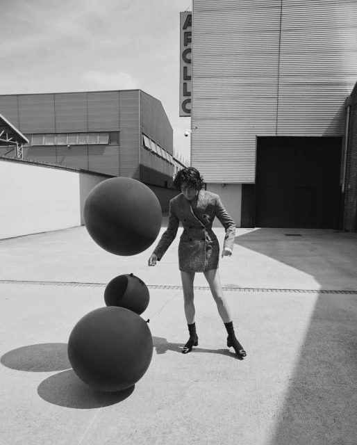 Black and white image of a woman in a short jacket and boots standing in front of modern buildings, holding black spheres.