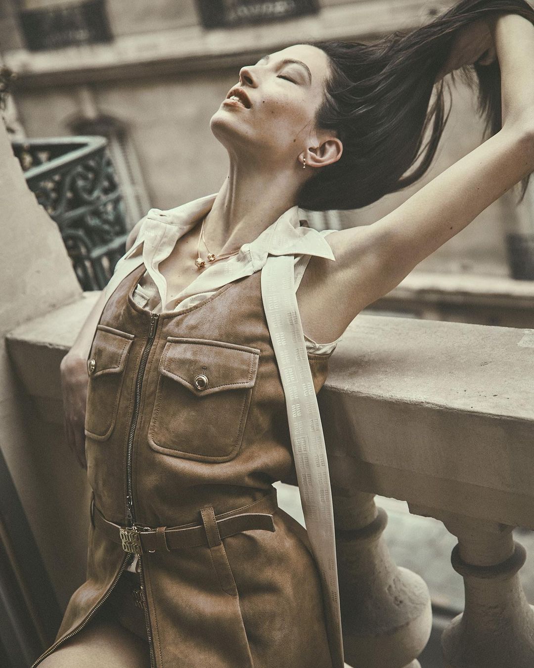 Stylish woman with long hair poses in a brown dress and white top on a balcony, bathed in soft light, creating a serene and elegant moment.