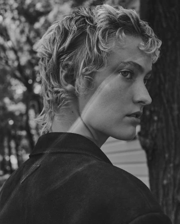 Monochrome portrait of a person with short curly hair in profile wearing a dark jacket outdoors against trees