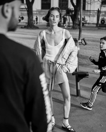 An adult woman in athleisure stands in a park with fitness equipment with two people in the background in a black and white photo.