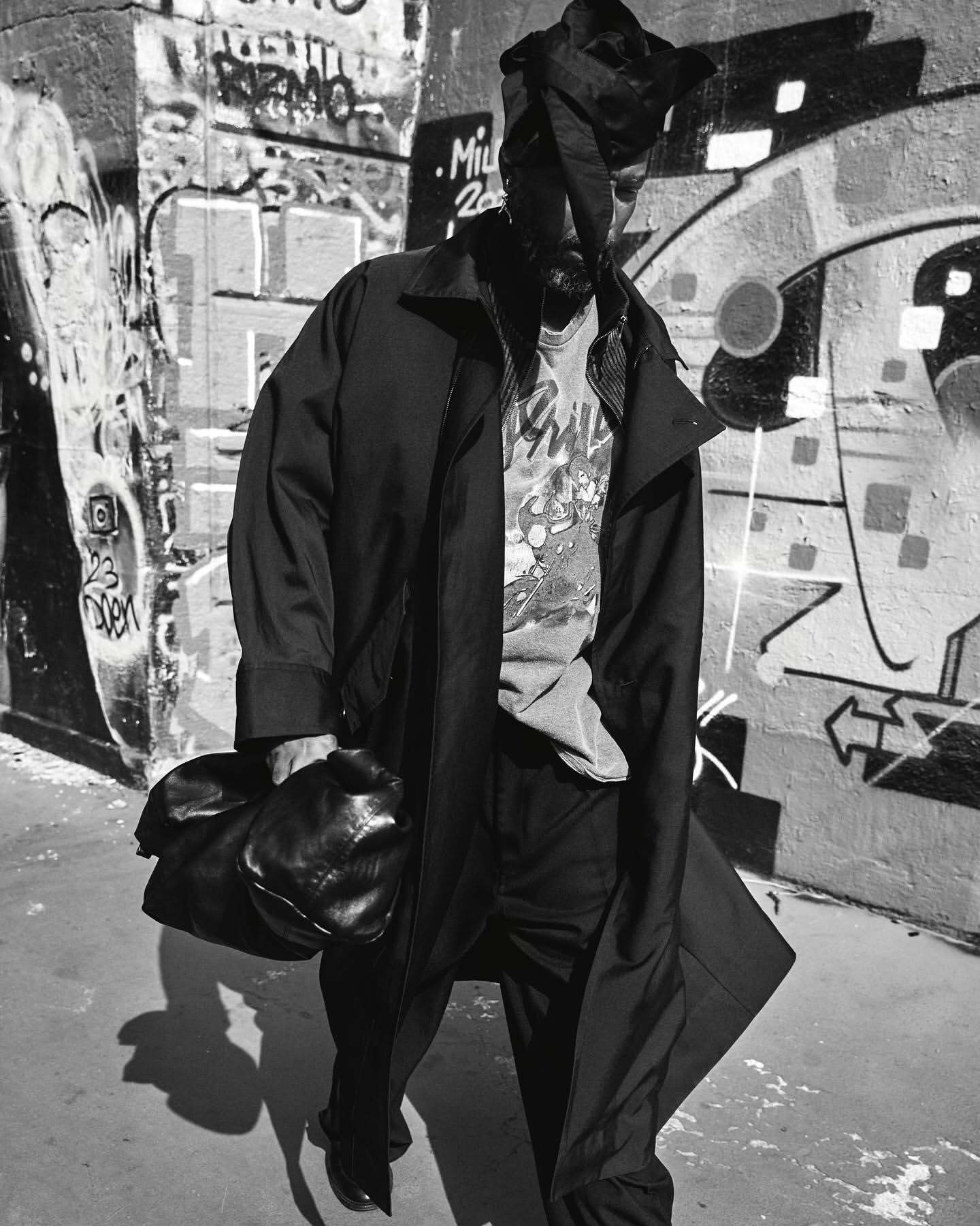 Black and white portrait of a man in a long coat and head wrap walking past a graffiti-covered wall, photographed by Stefano Galuzzi.