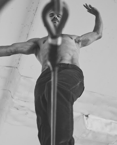 Low angle black and white view of a muscular adult male with a mustache, shirtless and wearing pants, inside an indoor environment.