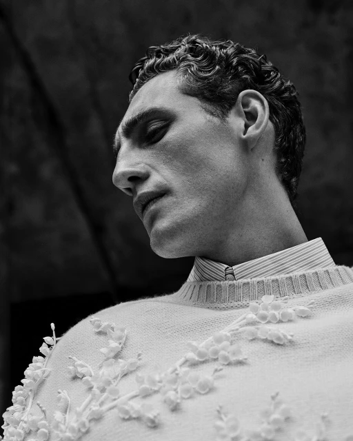 Monochrome portrait of an adult male in a light-colored, floral-patterned sweater, looking down with a neutral expression against a dark background.