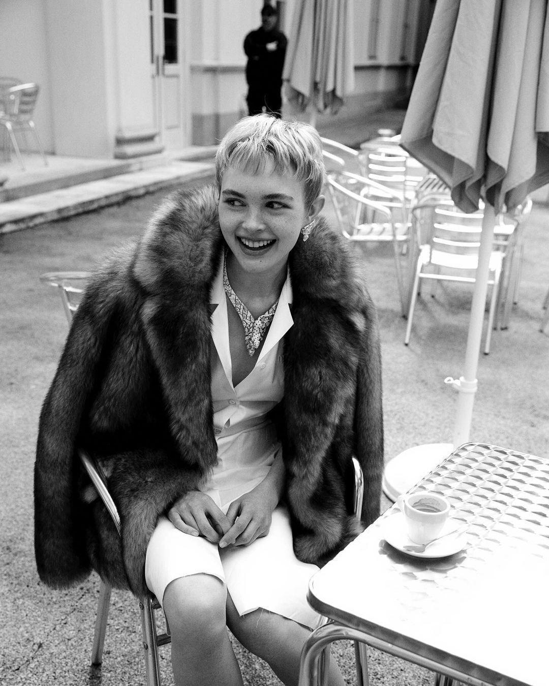 Woman with short hair in fur coat sits at table with coffee in black and white, smiling, in an outdoor cafe setting with neutral tones.