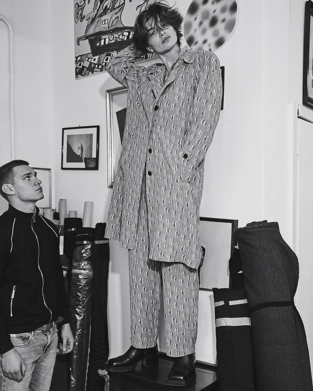 Monochrome shot of a male fashion model in patterned coat and pants, posing indoors next to another man in jeans and a jacket.