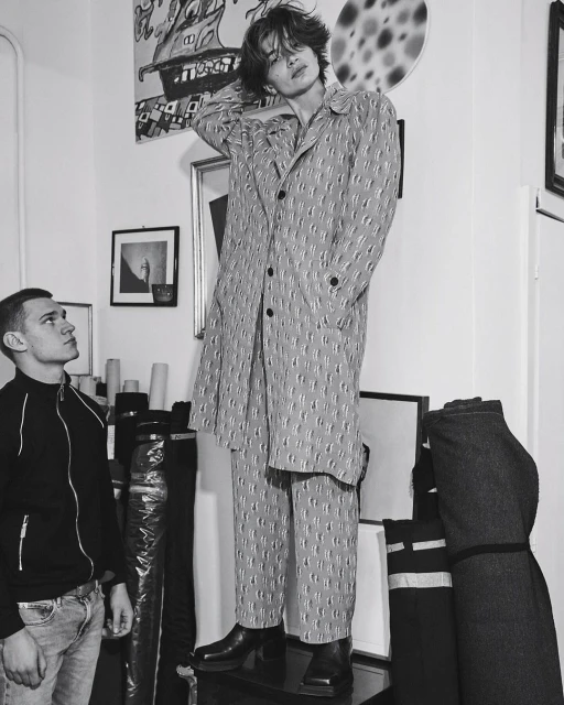 Monochrome shot of a male fashion model in patterned coat and pants, posing indoors next to another man in jeans and a jacket.