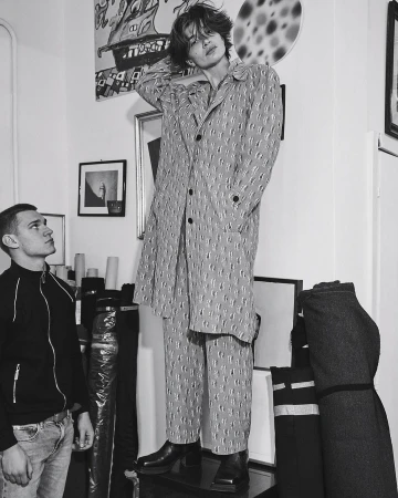 Monochrome shot of a male fashion model in patterned coat and pants, posing indoors next to another man in jeans and a jacket.
