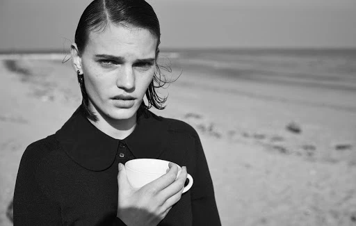 Black and white portrait of a woman on the beach holding a white cup, with a view of the sea and sky in the background.