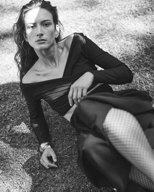 Monochrome portrait of a young adult woman lying on grass wearing a long-sleeved top, dress pants and fishnet stockings, eyes open.