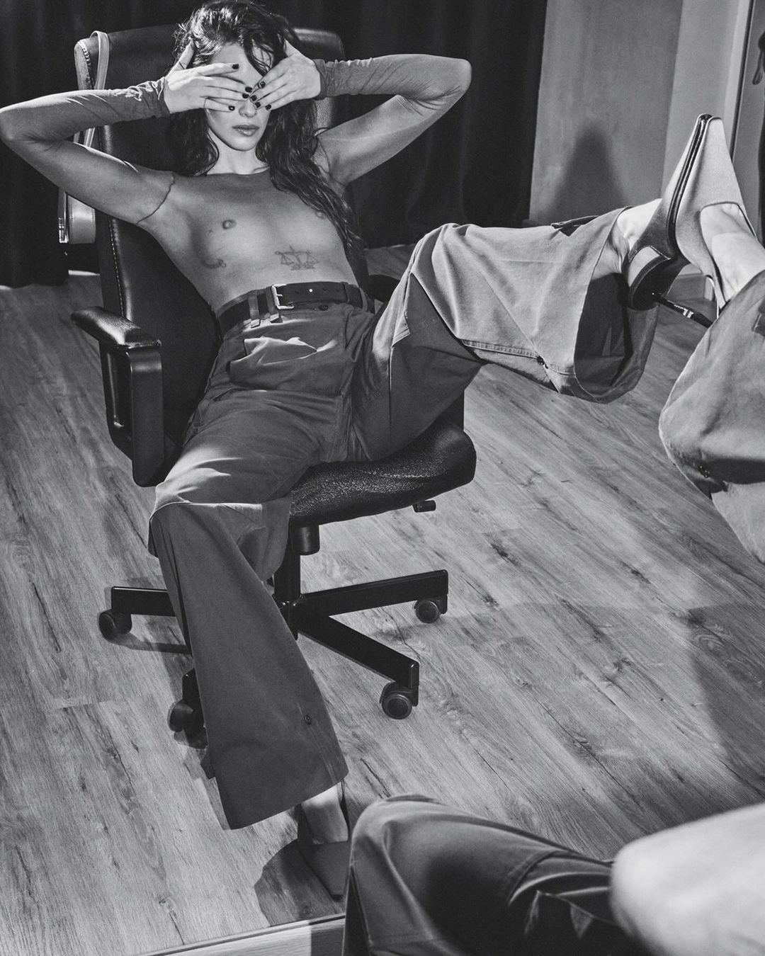 Monochrome portrait of a woman with long hair sitting in an office chair, covering her eyes with her hands, wearing wide leg pants and heels.