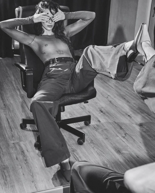 Monochrome portrait of a woman with long hair sitting in an office chair, covering her eyes with her hands, wearing wide leg pants and heels.