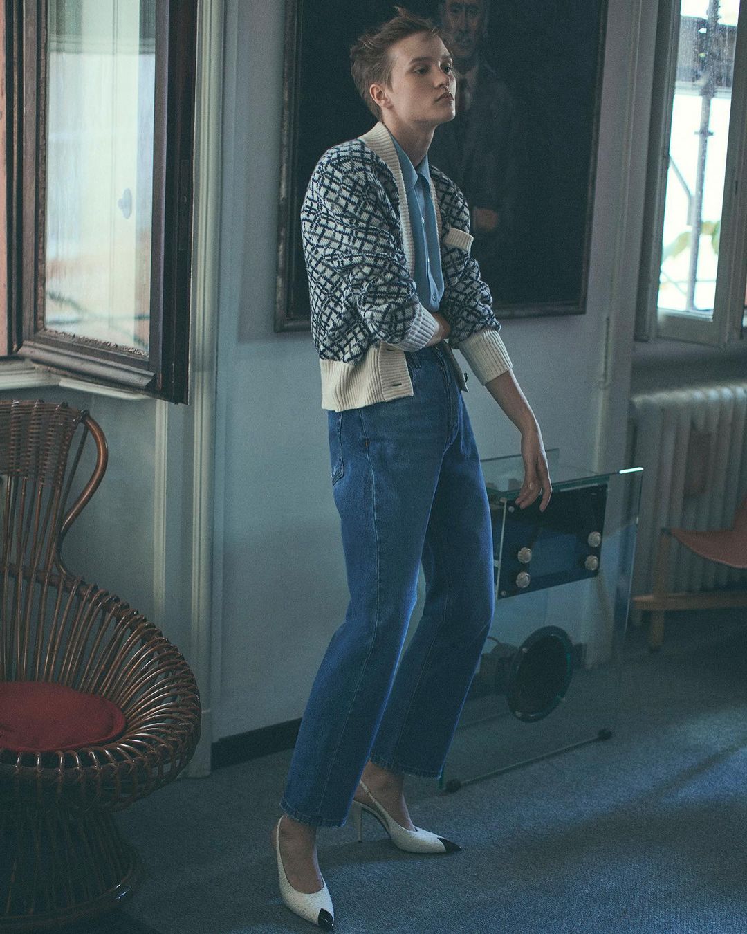 Fashion shot of woman indoors wearing cardigan, blue shirt, jeans, and pumps, posing in front of painting in a well-lit room.