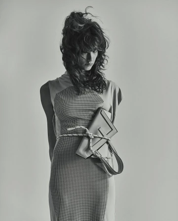 Model with voluminous dark hair wearing a patterned dress and holding a structured handbag tied with rope.