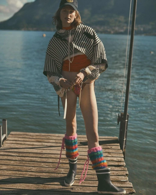 A woman poses on a wooden dock in a nautical-themed outfit. She wears a striped jacket, multicolored leg warmers, and a baseball cap.