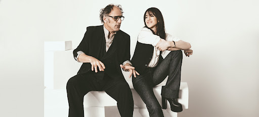 A casually posed photo features a man and a woman sitting on white blocks in front of a white backdrop, looking off in separate directions.