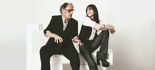A casually posed photo features a man and a woman sitting on white blocks in front of a white backdrop, looking off in separate directions.