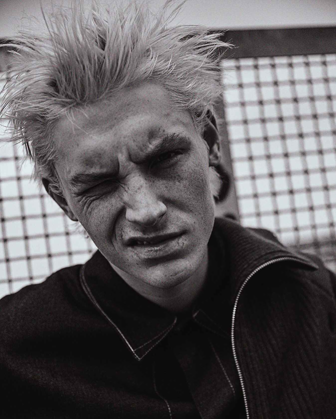 Monochrome portrait of a young adult with light hair, intense expression, wearing a dark textured jacket against a patterned background.