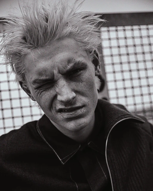 Monochrome portrait of a young adult with light hair, intense expression, wearing a dark textured jacket against a patterned background.