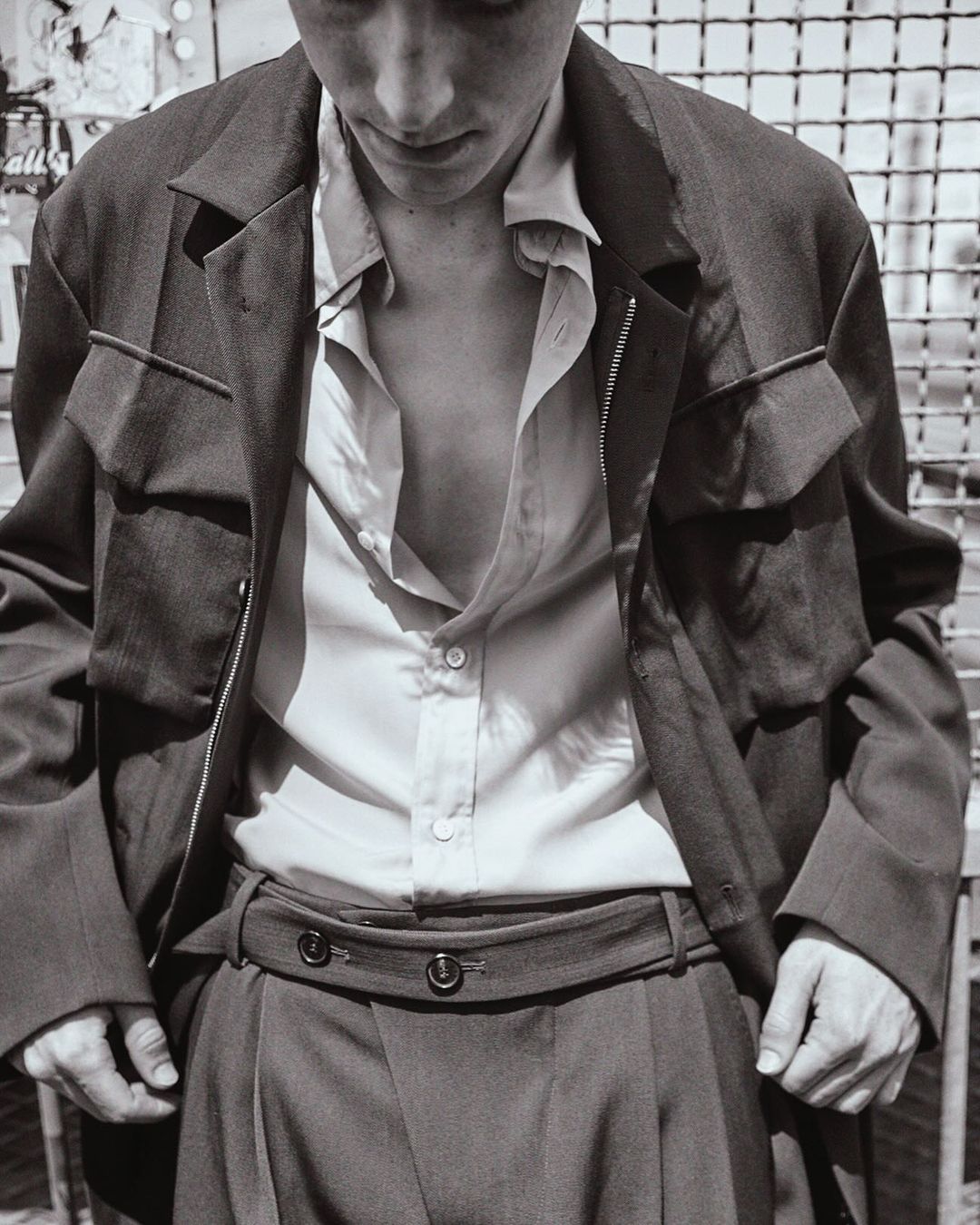 Monochromatic fashion portrait of a young adult man in an unbuttoned collared shirt, jacket, and high-waisted trousers against an urban backdrop.