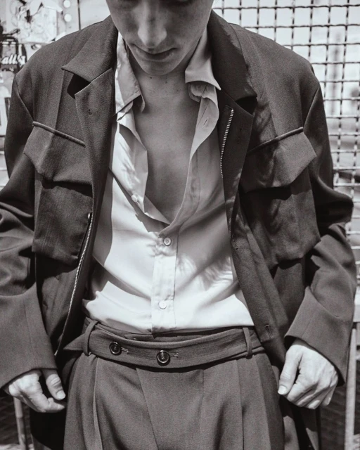 Monochromatic fashion portrait of a young adult man in an unbuttoned collared shirt, jacket, and high-waisted trousers against an urban backdrop.
