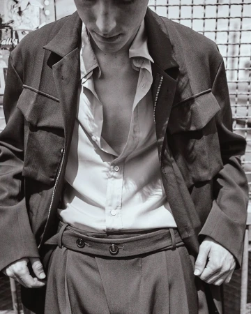 Man wearing a button-down shirt and utility jacket posing against a wire mesh fence background