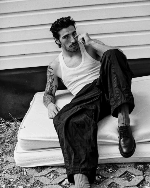 Monochrome portrait of a man with tattoos in a tank top and baggy pants, sitting on a mattress outdoors with a white wall backdrop.