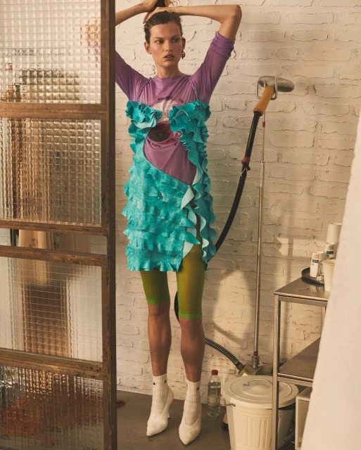 A woman in a bright turquoise dress and white boots poses indoors in a stylish fashion shot, looking away with her hands on her head.