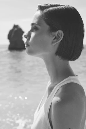 Black and white portrait of a woman with short dark hair in profile wearing a white tank top near the ocean.