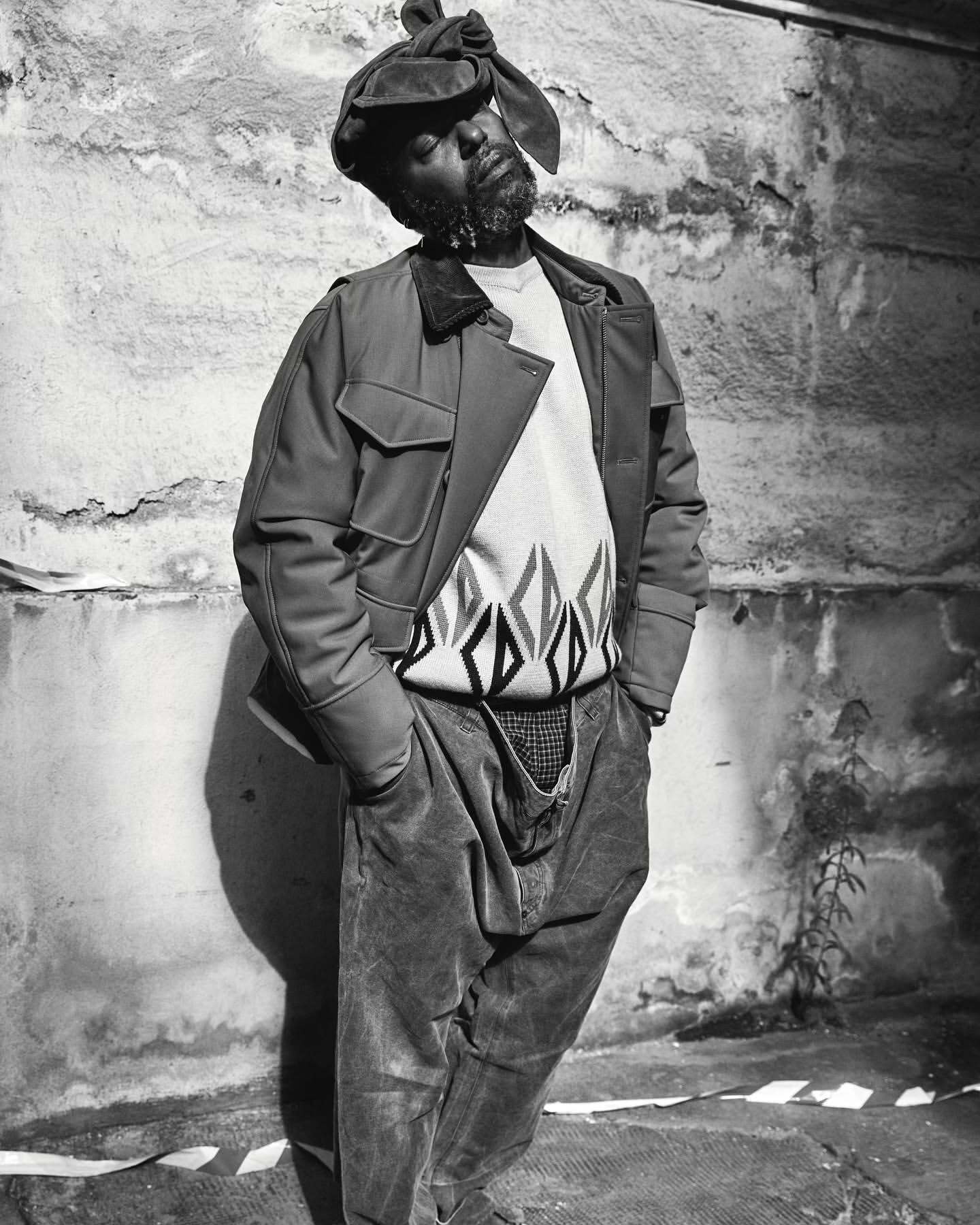 Monochrome portrait of an African American man in jacket and hat, standing against a textured wall, exuding a casual yet stylish vibe.