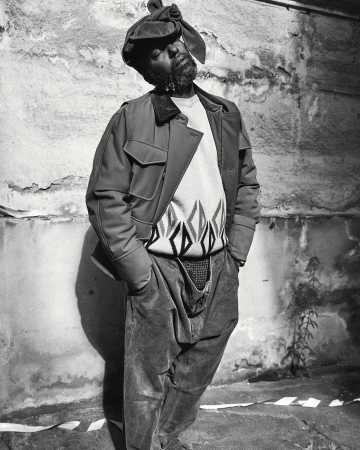 Monochrome portrait of an African American man in jacket and hat, standing against a textured wall, exuding a casual yet stylish vibe.