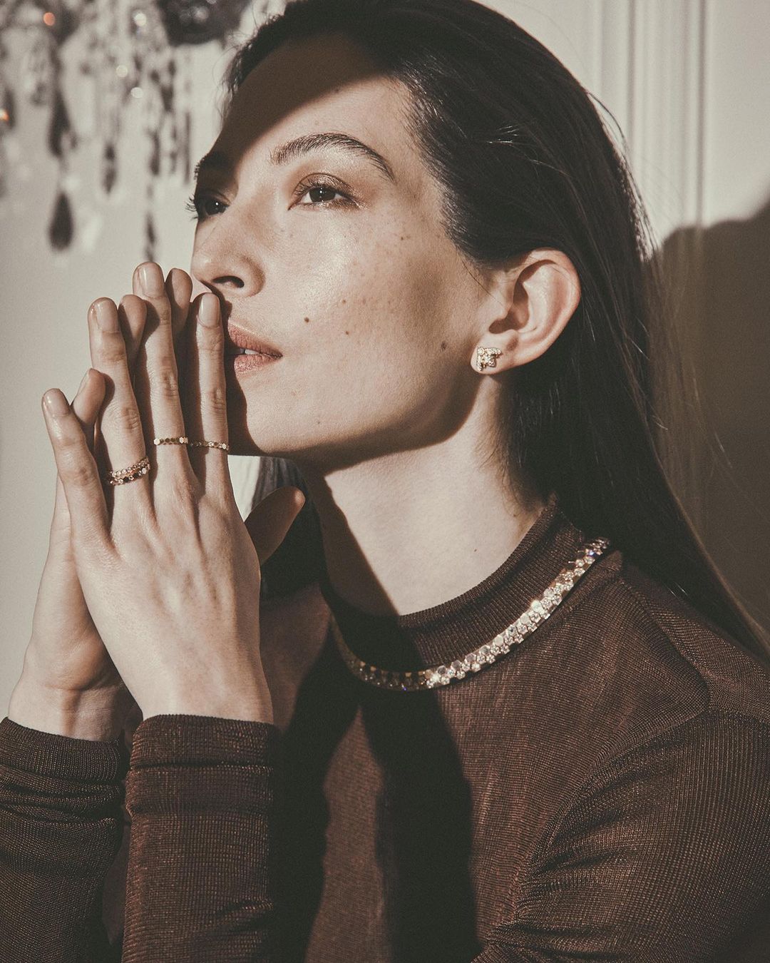 Elegant woman with her hands together, wearing jewelry, in a low-light indoor setting with a neutral background and a chandelier.