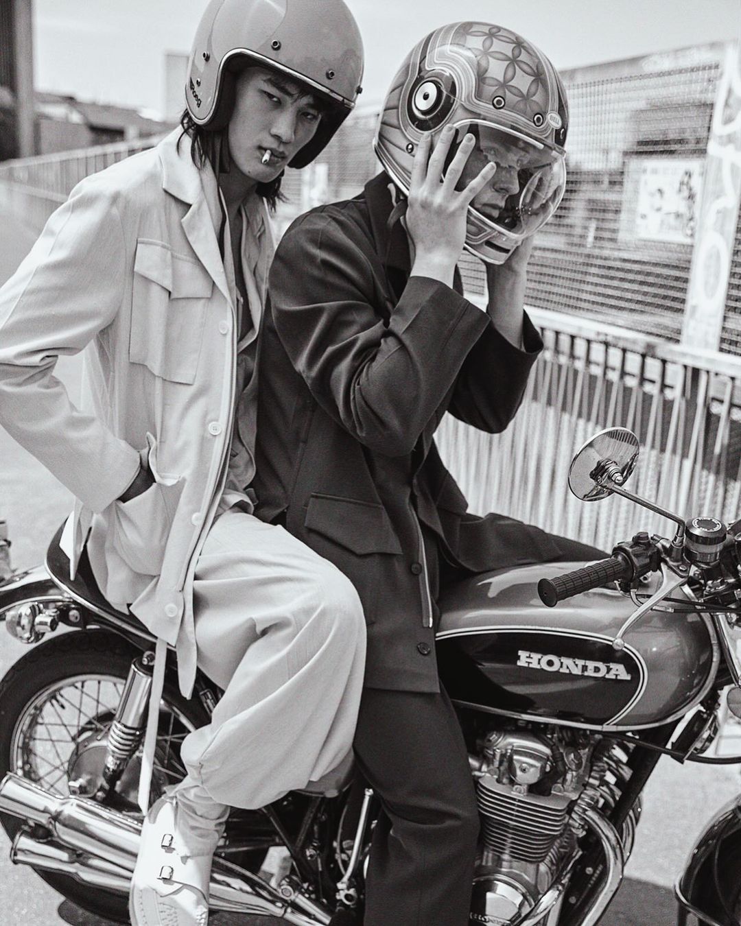 Two adult males pose on a Honda motorcycle wearing helmets in black and white. One man smokes. The background is urban.