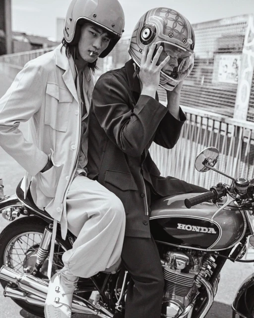Two adult males pose on a Honda motorcycle wearing helmets in black and white. One man smokes. The background is urban.