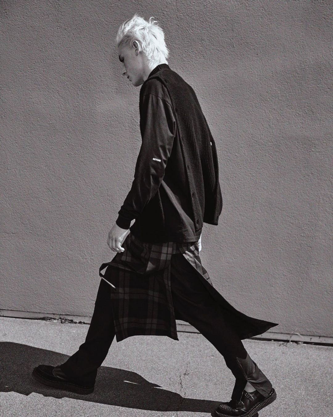Monochrome fashion shot of a blond male in a long-sleeved black shirt, pants, and a plaid skirt, walking against a textured gray wall.