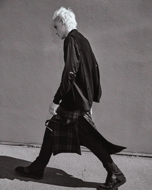 Monochrome fashion shot of a blond male in a long-sleeved black shirt, pants, and a plaid skirt, walking against a textured gray wall.
