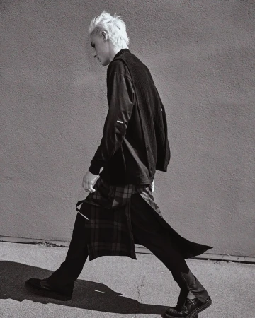 Monochrome fashion shot of a blond male in a long-sleeved black shirt, pants, and a plaid skirt, walking against a textured gray wall.
