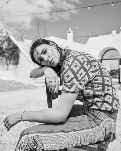 Black and white portrait of a young adult woman sitting on a chair in an outdoor setting with white buildings in the background.