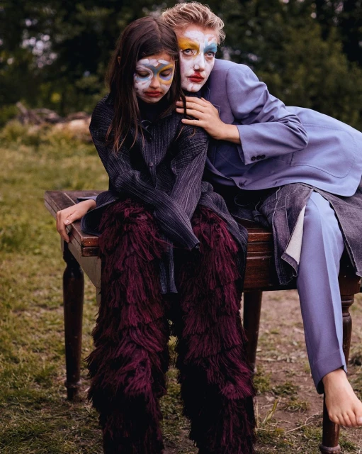 Two models, one a child, sit on a wooden table outdoors in an artistic fashion photograph with painted faces and fashion-forward clothing.