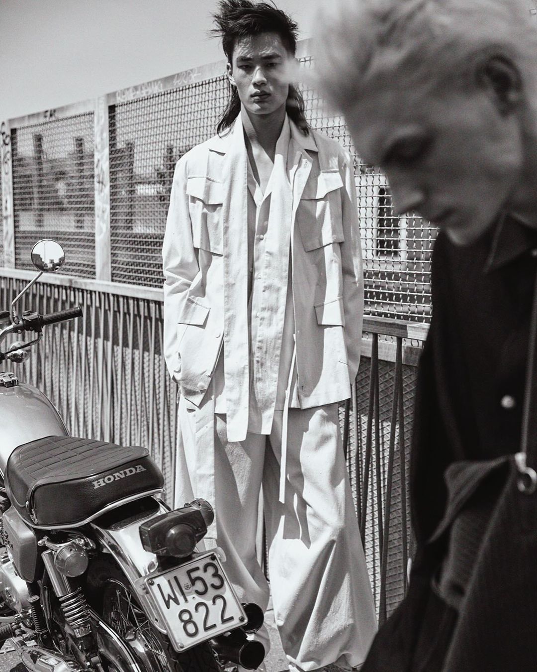 Monochrome outdoor fashion shot of two adult male models, one Asian, wearing casual white outfits next to a vintage Honda motorcycle.