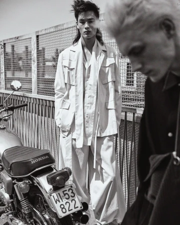Monochrome outdoor fashion shot of two adult male models, one Asian, wearing casual white outfits next to a vintage Honda motorcycle.