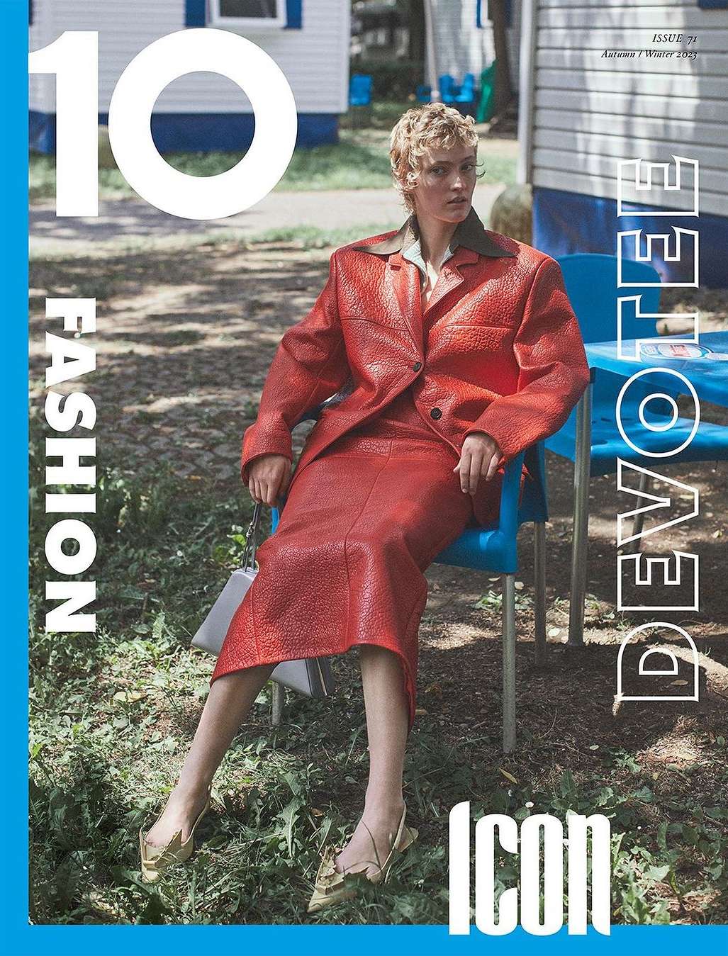 Fashion magazine cover featuring a model in a red leather suit sitting on a blue chair outdoors with a small house and greenery in the background.
