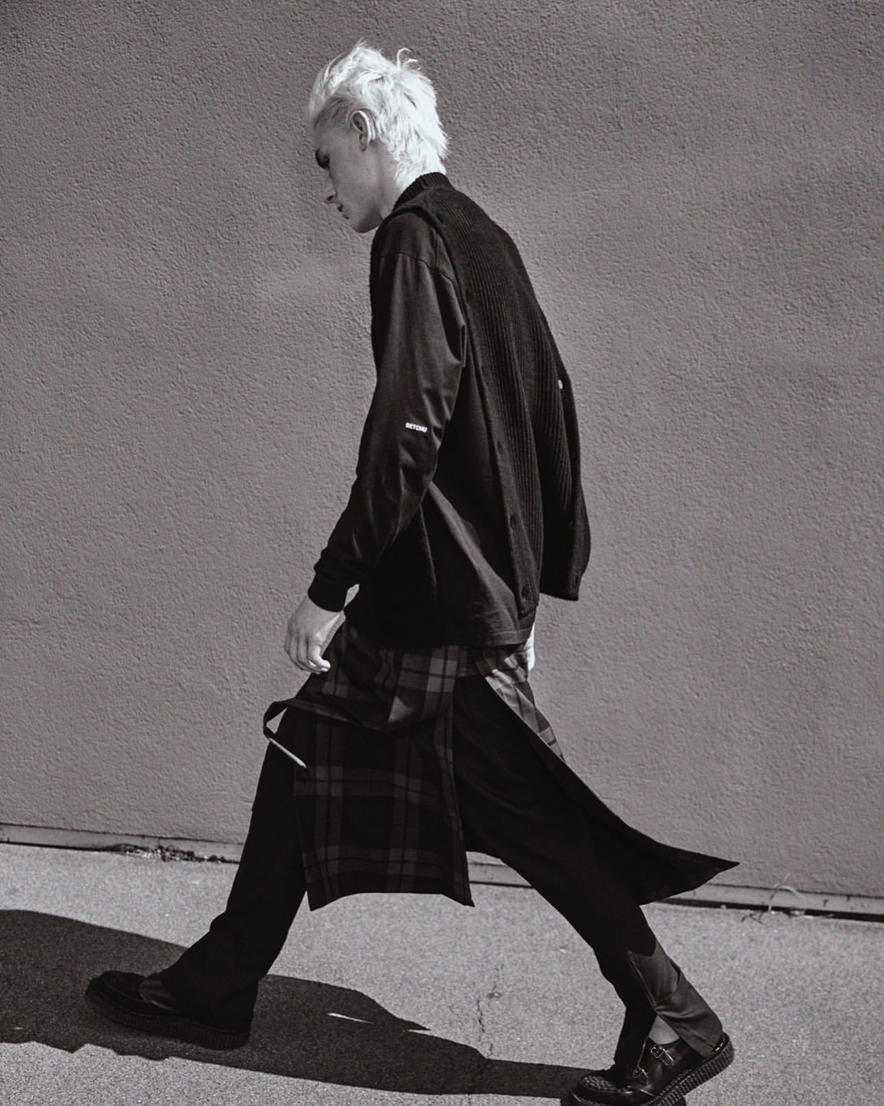 Monochrome portrait of a male model in stylish black clothing walking with a patterned kilt against a concrete wall backdrop in a fashion shoot.