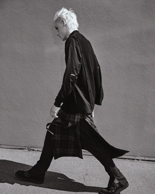 Monochrome portrait of a male model in stylish black clothing walking with a patterned kilt against a concrete wall backdrop in a fashion shoot.