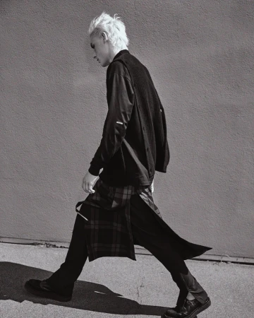 Monochrome portrait of a male model in stylish black clothing walking with a patterned kilt against a concrete wall backdrop in a fashion shoot.