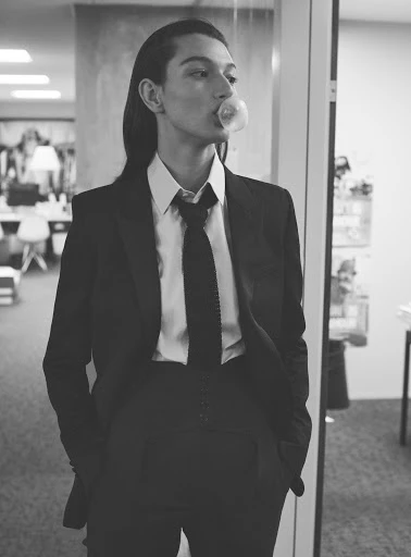 Black-and-white shot of a woman in a dark suit blowing a bubble with bubblegum in a modern office setting with natural light.