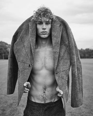 Monochrome portrait of a shirtless young man holding a heavy textured jacket draped over his shoulders outdoors.