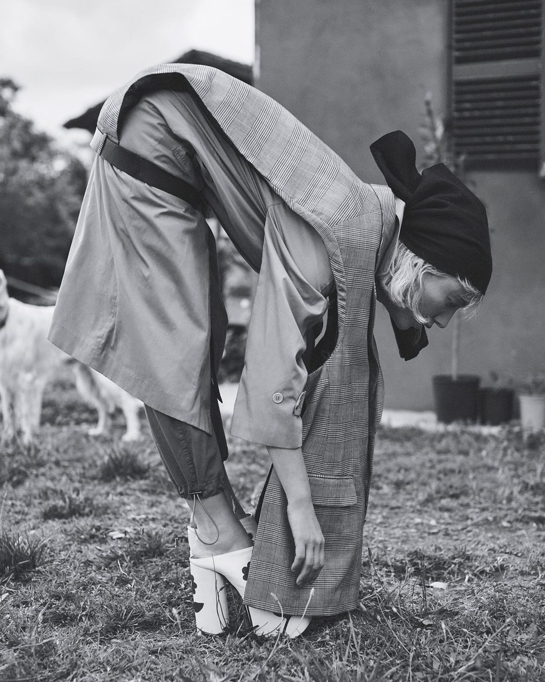Model bent over wearing patterned sleeveless coat, black headscarf, and platform mules in outdoor setting.