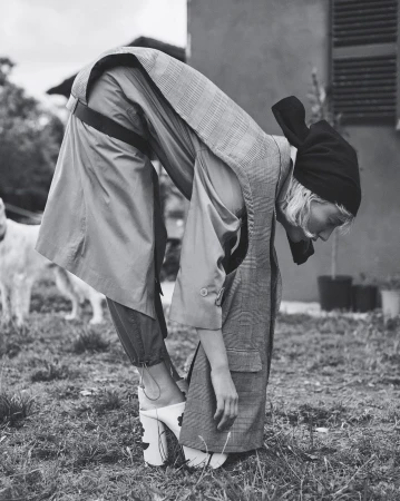 Model bent over wearing patterned sleeveless coat, black headscarf, and platform mules in outdoor setting.