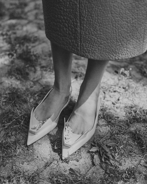 Low-angle shot of cream-colored pointed slingback heels on a woman standing on a dirt surface, below the knee, in a monochrome outdoor setting.
